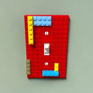 K&ouml;nnte beinhalten: Eine rote Lichtschalterabdeckung, verziert mit bunten Spielzeugbausteinen. Die Abdeckung hat einen Standard-Kippschalter und ist mit gelben, blauen und beigen Bl&ouml;cken verziert. Ein verspielter und kreativer Zusatz f&uuml;r jeden Raum.