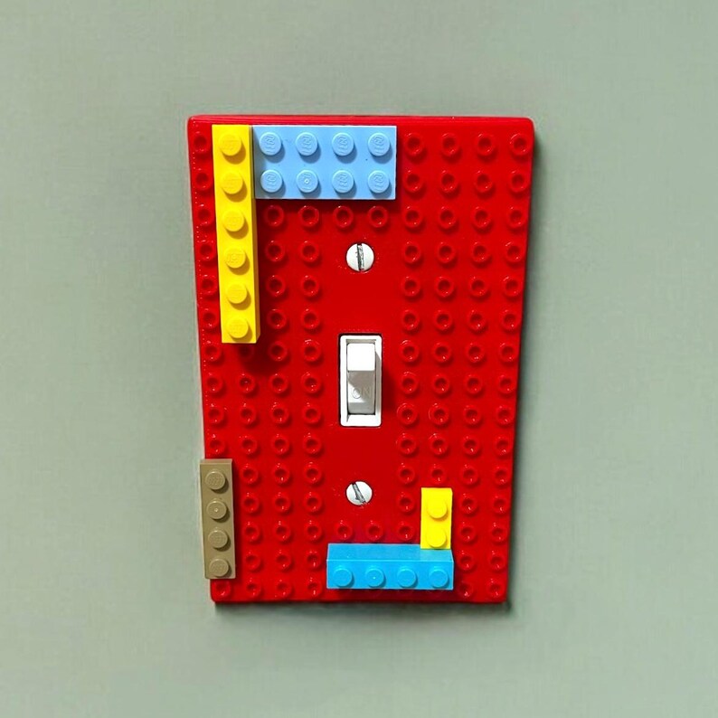 K&ouml;nnte beinhalten: Eine rote Lichtschalterabdeckung, verziert mit bunten Spielzeugbausteinen. Die Abdeckung hat einen Standard-Kippschalter und ist mit gelben, blauen und beigen Bl&ouml;cken verziert. Ein verspielter und kreativer Zusatz f&uuml;r jeden Raum.