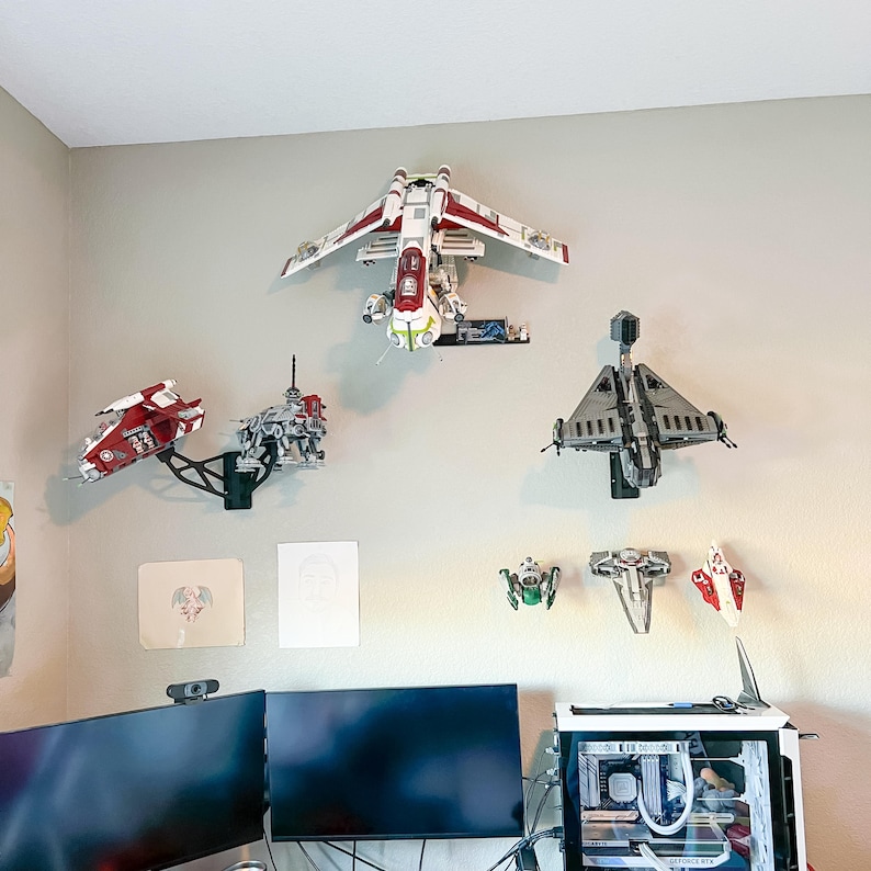 May include: A collection of Star Wars LEGO models mounted on a light gray wall. The models include various spacecraft and vehicles in red, white, gray, and green. A computer setup with dual monitors is visible below.