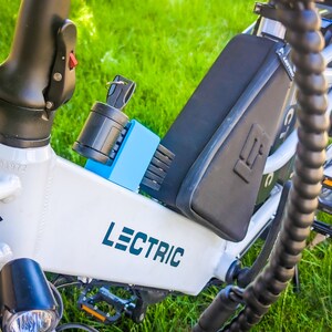 Pu&ograve; includere: Telaio di bicicletta elettrica Lectric bianco con componenti neri, un pacco batteria nero e un lucchetto blu. La bici ha la scritta "LECTRIC" in turchese. Lo sfondo &egrave; erba verde.