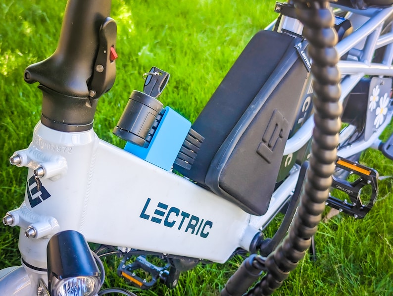 Pu&ograve; includere: Telaio di bicicletta elettrica Lectric bianco con componenti neri, un pacco batteria nero e un lucchetto blu. La bici ha la scritta "LECTRIC" in turchese. Lo sfondo &egrave; erba verde.