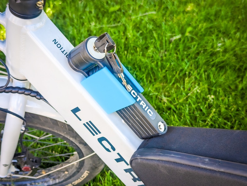 Pu&ograve; includere: Primo piano di un telaio di bicicletta elettrica bianco con un lucchetto pieghevole nero e blu attaccato. Il lucchetto &egrave; assicurato con le chiavi. La parola "LECTRIC" &egrave; stampata sul telaio e sul lucchetto.