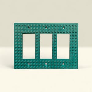 K&ouml;nnte beinhalten: Eine teal-farbene Lichtschalterplatte mit drei rechteckigen &Ouml;ffnungen. Die Platte hat eine strukturierte Oberfl&auml;che mit kleinen, kreisf&ouml;rmigen Noppen, die an ein Bausteindesign erinnern. Die rechteckige Platte hat eine klare, moderne &Auml;sthetik.
