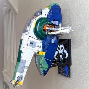 Puede incluir: Maqueta Lego de una nave espacial blanca, verde y azul, montada en una pared. El modelo presenta detalles intrincados y se exhibe con una placa negra con un emblema blanco y texto. La nave espacial es una réplica detallada.