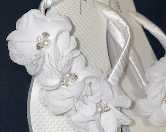 May include: White flip flops with white satin straps and white fabric flowers with pearl and rhinestone embellishments. The flip flops have the text "OLD NAVY" printed on the straps.
