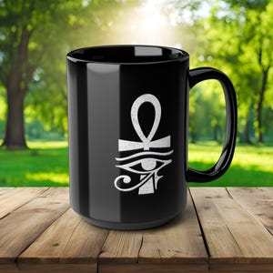 May include: A black ceramic mug featuring a white Ankh symbol and the Eye of Horus. The mug has a large handle and is set on a wooden surface with a blurred green background.