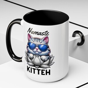 May include: White ceramic mug with a black handle and interior. The mug features a cartoon cat wearing sunglasses and sitting on a lotus flower. The words "Namaste" and "Kitteh" are printed on the mug.