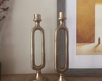 May include: Two gold metal candle holders with a unique, modern design. The holders have a rectangular base and a tall, slender body with an open oval shape in the middle.