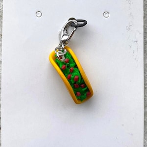 May include: A miniature taco charm with a yellow shell, filled with green lettuce and red tomatoes. The charm is attached to a silver-colored clasp and a black cord, perfect for a keychain or bracelet.