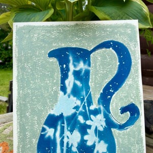 May include: A rectangular art print featuring a cyanotype-style blue pitcher silhouette against a textured light green background. The pitcher displays white floral and botanical negative imprints within its deep blue body, showcasing a curved handle.