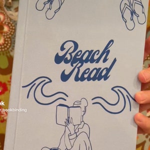 May include: A light blue book cover with the words "Beach Read" in a dark blue script. The cover features dark blue illustrations of flip-flops, waves, and a person reading. A beige ribbon is attached to the top.