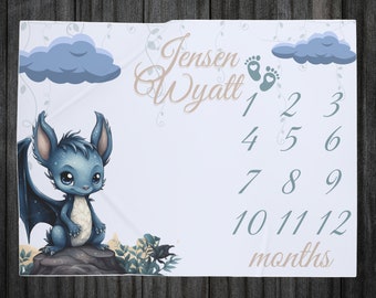 May include: A baby milestone blanket with a blue and white color scheme. It features a cute blue dragon illustration, clouds, and vines. The blanket has a number chart for tracking a baby's growth from 1 to 12 months. The text "Jensen Wyatt" is written in a cursive font at the top of the blanket.