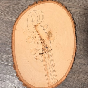 May include: A wood slice with a woodburned image of a cello neck and scroll. The wood is light brown with a natural grain pattern.