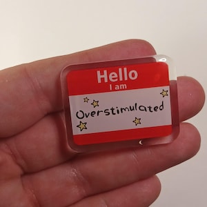 May include: A rectangular acrylic pin featuring a red and white name tag design. The top red section reads "Hello I am" in white text. The white middle section features the word "Overstimulated" in black script with four small yellow stars.