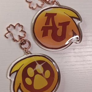 May include: Two clear acrylic keyrings with copper-coloured fixings. One keyring displays the letters "AJ" in a brown font on a yellow and orange background. The other has a paw print design in a similar colour scheme.