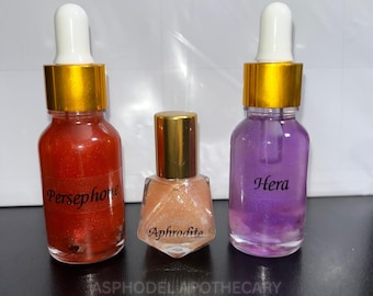 May include: Three glass bottles with gold caps. The left bottle contains orange glitter liquid labelled Persephone. The centre bottle is diamond-shaped with peach glitter liquid labelled Aphrodite. The right bottle contains purple glitter liquid labelled Hera.