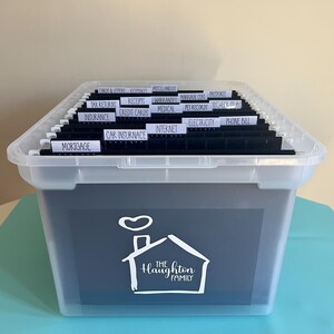 May include: A clear plastic file box with a lid, filled with hanging file folders. The folders are labeled with categories like "Mortgage," "Insurance," and "Credit Cards." The box has a dark blue front with a white house graphic and the text "The Haughton Family."