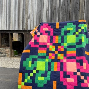 Könnte beinhalten: Eine große Steppdecke mit einem pixeligen geometrischen Muster aus dunkel marineblauen, neongrünen, leuchtend pinken und orangefarbenen Stoffquadraten. Das Design ist symmetrisch und besteht aus abwechselnden Farbblöcken in einem Raster.
