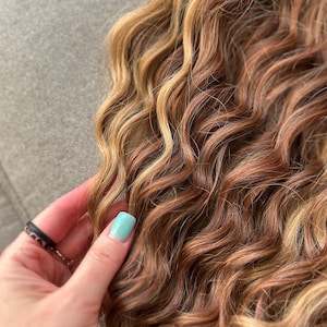 May include: Close-up of wavy, multi-toned hair extensions. The hair features a blend of light blonde, golden brown, and reddish-brown hues. The texture is curly, with each strand showing a natural wave pattern. The hair is soft and appears to be high-quality.