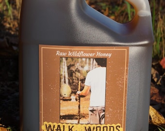 May include: A large, dark gray plastic jug with a white cap and handle, filled with raw wildflower honey. The label features a brown background with the text "Raw Wildflower Honey" and an image of a person walking in the woods. The label also reads "Walk in the Woods Apiary."
