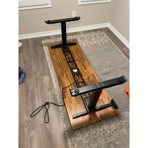 May include: A black standing desk frame with a wooden top. The frame has two black metal legs and a black metal crossbar. The wooden top is a light brown colour and has a wood grain pattern.