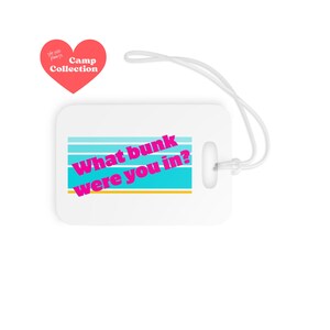 May include: White luggage tag with the text "What bunk were you in?" in pink and blue. The tag has a blue and white striped design and a yellow stripe at the bottom. A red heart-shaped logo with the words "Camp Collection" is in the upper left corner.