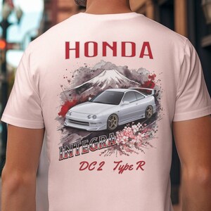 Puede incluir: Una camiseta blanca con un estampado gráfico de un Honda Integra DC2 Type R blanco frente al monte Fuji con flores de cerezo. El texto "HONDA" e "INTEGRA DC2 Type R" está impreso en rojo en la camiseta.