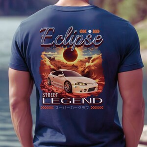 Puede incluir: Una camiseta azul marino con un gráfico de un coche blanco y el texto "Eclipse" y "Street Legend" en una fuente de estilo retro. El coche está conduciendo por un paisaje desértico con un eclipse solar en el fondo. El texto "スーパーカークラブ" también está impreso en la camiseta.