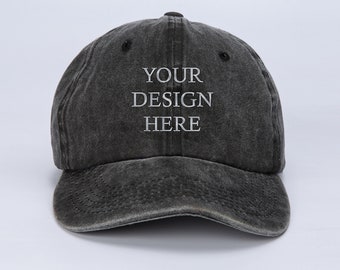 Op de afbeelding: Een zwarte, verweerde, katoenen baseballpet met een wit geborduurd ontwerp dat "YOUR DESIGN HERE" zegt.