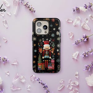 Może przedstawiać: Czarne etui na telefon z świątecznym wzorem dziadka do orzechów, otoczone płatkami śniegu, choinkami i pudełkami z prezentami. Na lawendowym tle z gałązkami lawendy i kryształami. Widoczny tekst "Lavender Love".