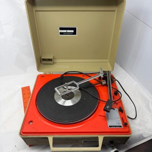May include: A beige and bright orange General Electric portable record player with a black turntable mat, a metal record spindle arm, and a black power cord. An orange ruler is visible on the left, showing a length of 10 inches.
