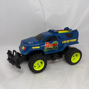 May include: A blue Tyco Python remote control truck featuring bright neon yellow wheels, a black front bumper, and a long antenna. The body displays the text Python, Tyco, and 6.0V Jet Turbo in yellow and red graphics with a green snake illustration.