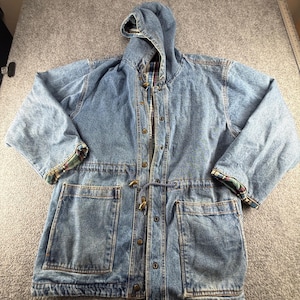 May include: A light blue denim hooded jacket featuring a front button closure, a drawstring waist with toggle fasteners, and two large patch pockets. The interior and sleeve cuffs are lined with a multi-colored plaid fabric pattern.