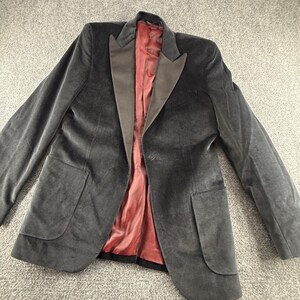 May include: A dark charcoal gray velvet blazer featuring a single-button closure, wide satin lapels, and two large patch pockets on the front. The interior of the jacket is fully lined with a smooth, solid deep red fabric throughout the body.