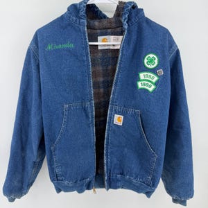 Pode incluir: Casaco de ganga azul com capuz e fecho de correr. O casaco tem um nome bordado a verde, patches com os anos 1998 e 1999 e um emblema de trevo de quatro folhas. O casaco é forrado com um padrão xadrez.