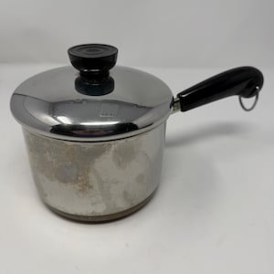 May include: A small, stainless steel saucepan with a lid and a black handle. The lid has a black knob. The handle has a loop at the end. The pan shows signs of wear.
