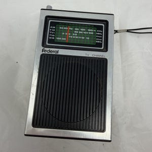 以下が含まれることがあります： 銀色の金属製フレームとスピーカーグリル付きの黒いフロントパネルを備えた長方形のポータブルラジオ。ディスプレイにはFederalのブランド名、AM、FM、TV、WBバンド用の緑色の周波数スケール、およびテレビチャンネルセレクターが表示されています。