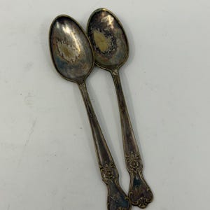 May include: Two tarnished silver-toned metal spoons with an oval bowl and a decorative handle featuring an embossed floral motif at the base. The metal surface shows significant oxidation and dark patina throughout the stems and bowls.