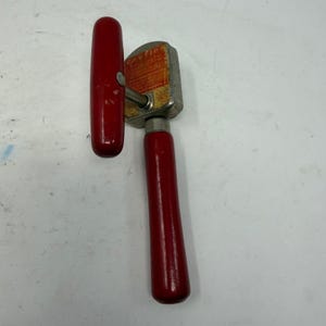 May include: A vintage manual can opener featuring two glossy red wooden handles and a metallic head. The central metal component displays a rectangular orange label with the word CAUTION printed in bold black letters at the top of the text block.