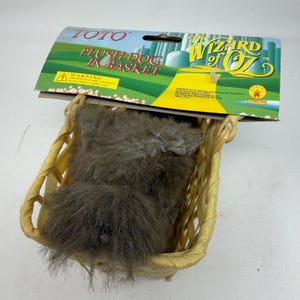 May include: A plush dog toy in a woven basket, with a cardboard header featuring the text "TOTO PLUSH DOG IN BASKET" and "The Wizard of Oz" logo. The dog is gray and fluffy.