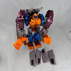 May include: A robotic action figure with a gorilla-like torso, orange chest, and blue metallic accents. It features large orange claw hands, purple shoulder armour, and two grey cannons on its back. Four separate grey rectangular panels lie in front.