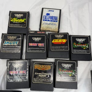 Peut inclure: Un ensemble de neuf cartouches de jeux vidéo rectangulaires noires pour le système ColecoVision, incluant des titres comme Looping, California Games, Smurf, Donkey Kong, Gorf, Venture et Buck Rogers, avec des étiquettes imprimées.