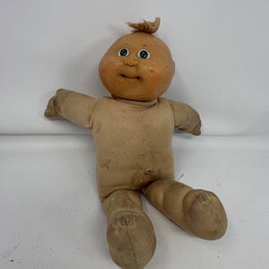 May include: A vintage fabric doll with a round, tan-colored head, a small tuft of light brown hair on top, and green eyes. The doll has a soft, beige cloth body with visible signs of wear and discoloration, featuring a simple stitched navel.