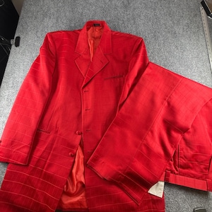 May include: A bright red suit set consisting of a long-sleeved blazer and matching trousers. The blazer features a notched lapel, a button-front closure, and a subtle grid pattern throughout. The fabric has a slight sheen and a red lining.