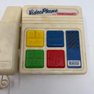May include: A vintage off-white toy VideoPhone from VideoSmarts. The phone has a handset and a base with colored buttons labeled True, False, Yes, and No. The base also features a blue speaker and the VTech logo.