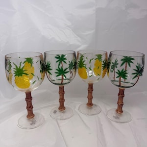 Könnte beinhalten: Ein Set aus vier klaren Weingläsern mit bemalten tropischen Motiven. Zwei Gläser zeigen gelbe Ananas mit grünen Blättern, während zwei weitere grüne Palmen darstellen. Jeder Stiel ist so strukturiert, dass er braunen Bambusrohren ähnelt.