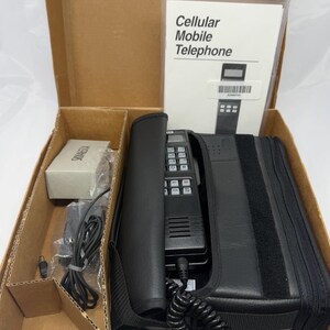 May include: A vintage black cellular mobile telephone with a numeric keypad and coiled cord, housed in a black carrying case. The set includes a power adapter, a small box, and an instruction manual titled Cellular Mobile Telephone.
