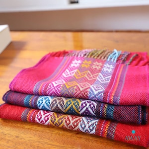 May include: A stack of four colourful woven scarves with geometric patterns in shades of pink, red, blue, yellow, and white. The scarves have fringed edges and are folded neatly on a wooden surface.