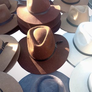Op de afbeelding: Een assortiment vilten hoeden in verschillende tinten bruin, beige en blauw. Elke hoed heeft een afgeronde kroon en een brede rand, met een donkere leren band rond de basis. De hoeden zijn dicht bij elkaar geplaatst en tonen hun verschillende kleuren en stijlen.
