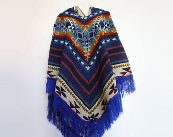 May include: A blue poncho featuring a vibrant, multi-colored geometric pattern with shades of orange, yellow, and white. The garment has a V-shaped neckline and is finished with long, deep blue fringe along the bottom hemline.